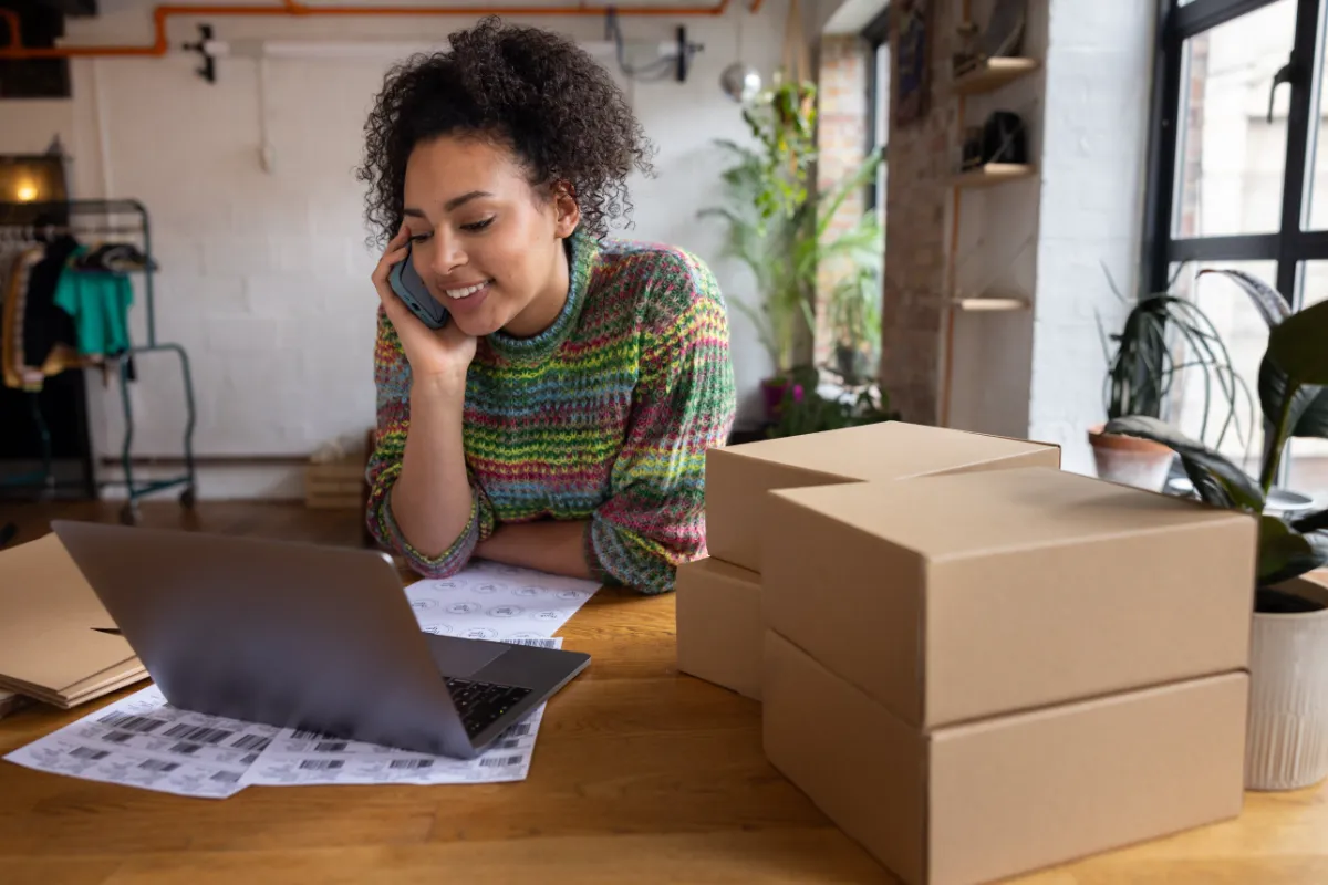 Online shop owner on the phone at her laptop with parcels ready to ship